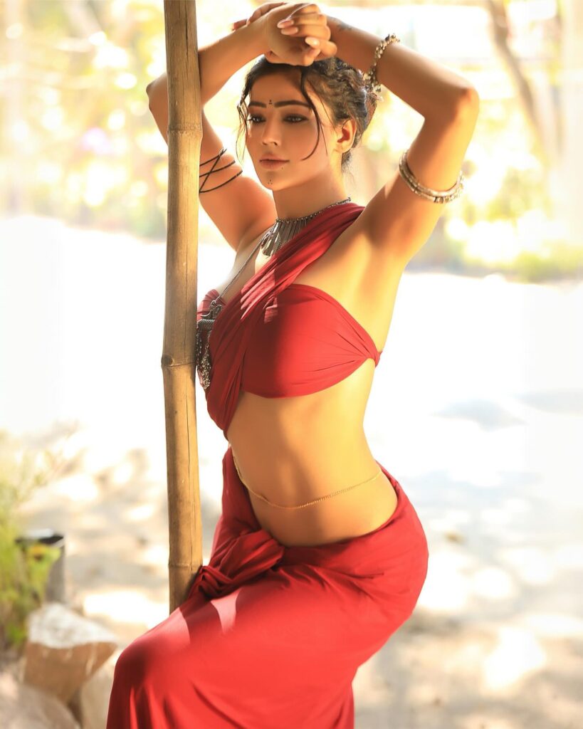 Sonia bansal in a red sari. She is standing next to a wooden pole and looking at the camera. The woman has long black hair and brown eyes. She is wearing a silver necklace and earrings.